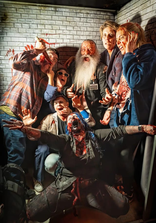 Seven zombies crammed into a photo booth against a white-brick backdrop flecked with blood: one brandishing a kitchen knife, one in dark glasses, a long-bearded walker in a ZOMBIE BUSINESS lanyard, and a blue-haired figure kneeling at the front with their arms flung wide.
