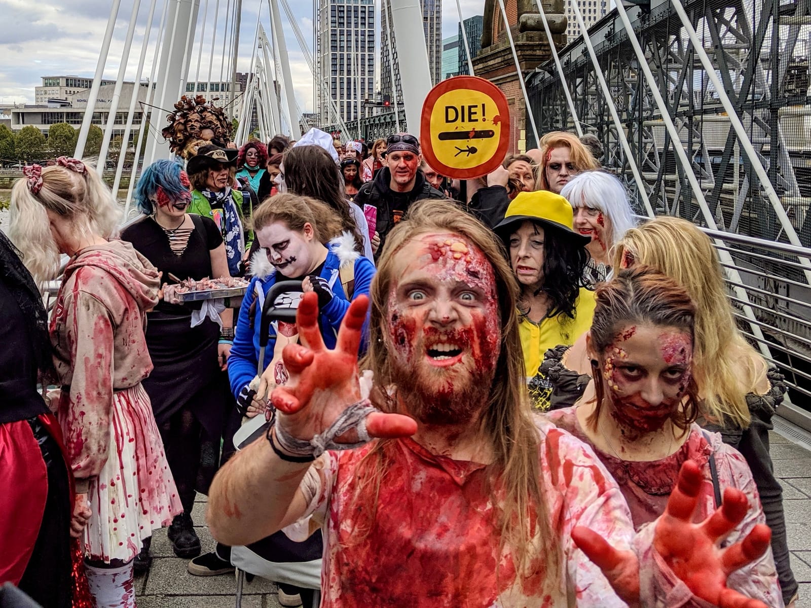 A zombie with bloodied outstretched hands lunges towards the camera on Golden Jubilee Bridge, a crowd of costumed zombies and a hand-painted DIE! road sign behind him.