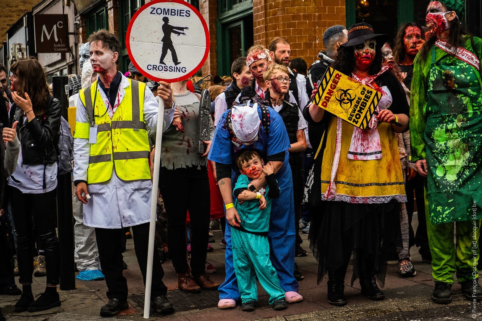 Zombies of all ages wait at the kerb: a steward holding a Zombies Crossing sign, a parent in scrubs with a small child in costume, and a zombie holding a yellow “Danger: beware of zombies” sign.