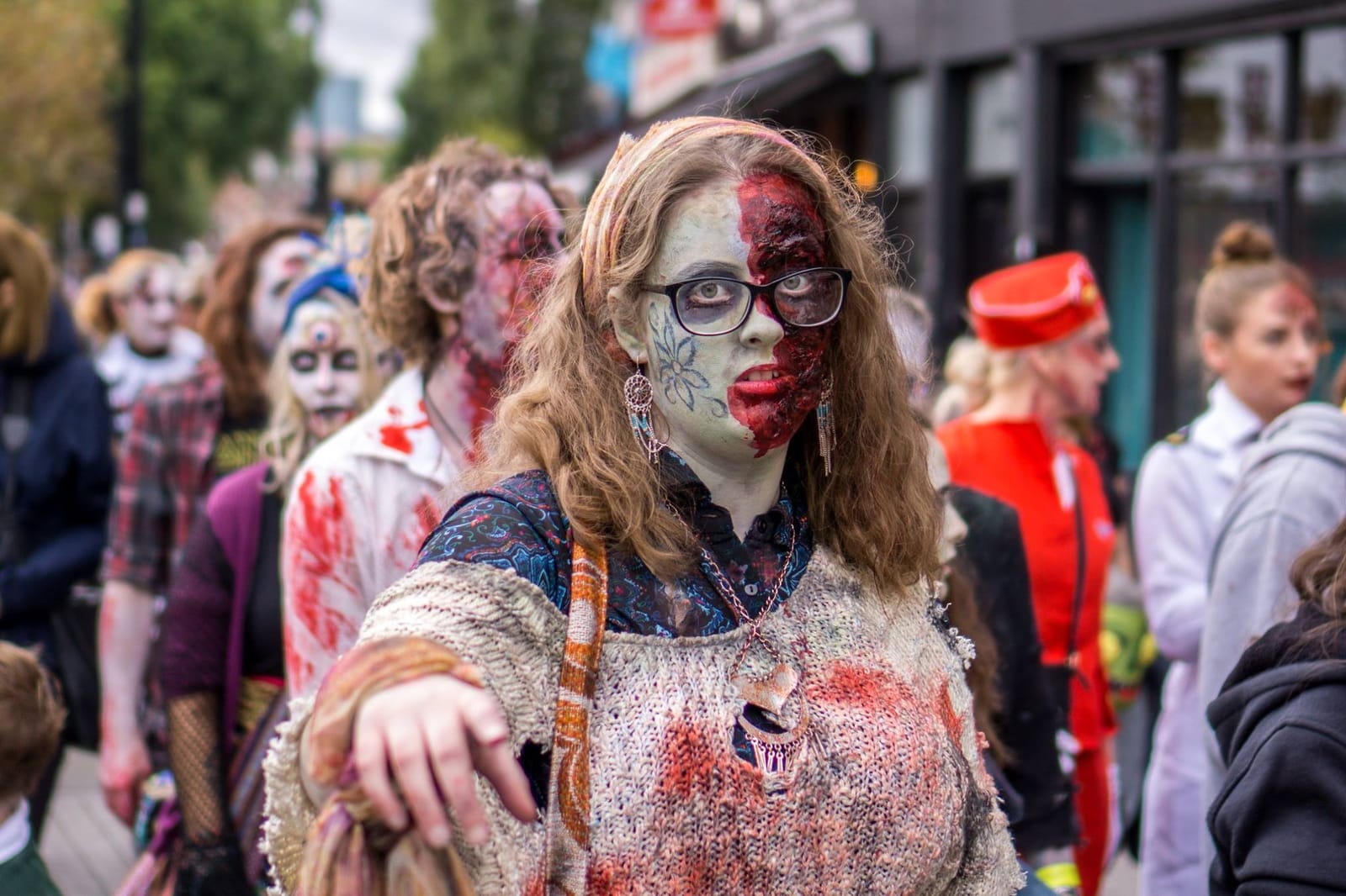 A zombie in glasses, half her face made up as raw flesh, reaches towards the camera with more zombies out of focus behind her.