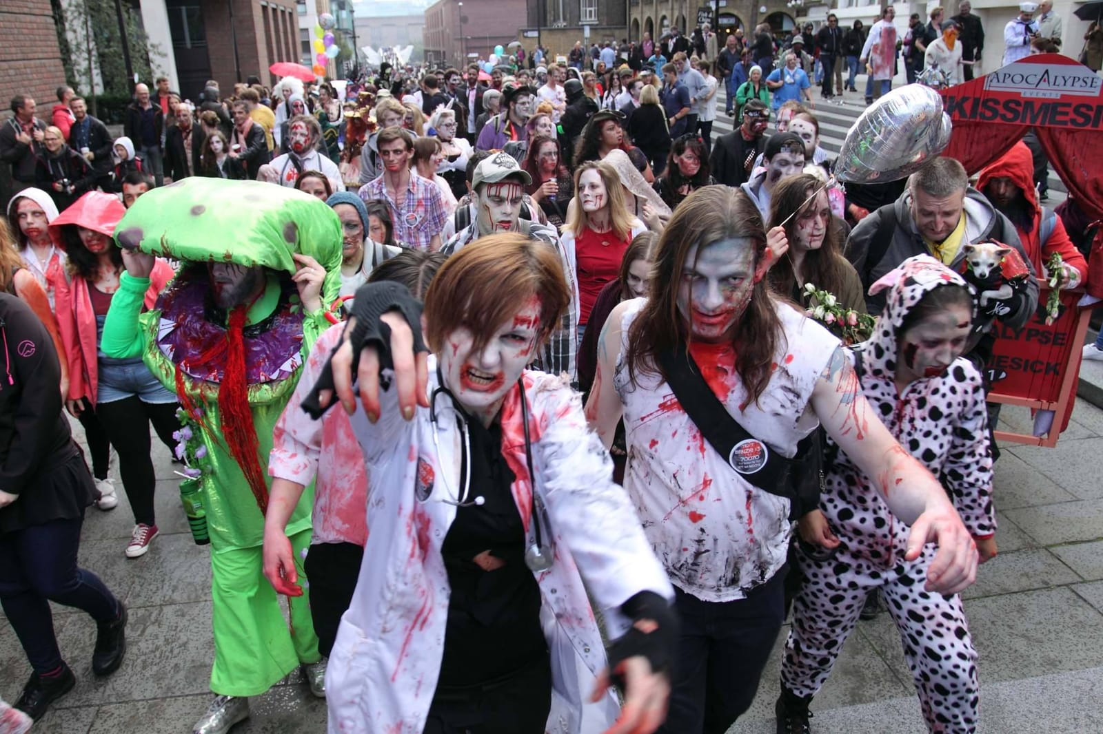 Zombies shamble past a sponsor’s red kissing-booth tent, one of them carrying a small dog and another holding a bunch of flowers.
