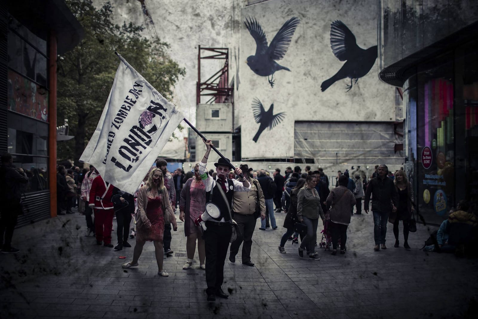A zombie in a bowler hat carries a megaphone and the World Zombie Day: London banner through Leicester Square, with the walk following behind.