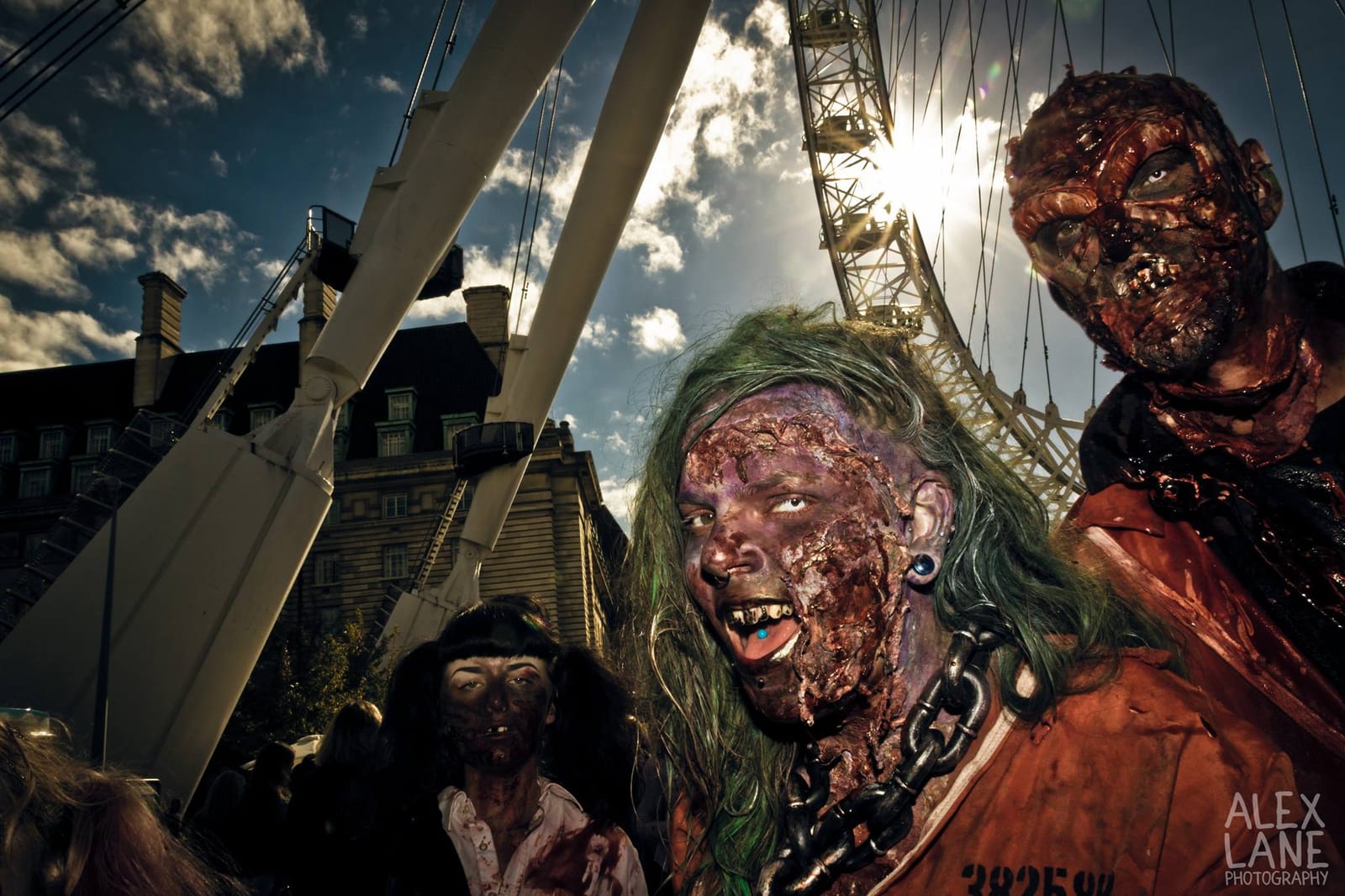 Two zombies in heavy prosthetic make-up pose beneath the London Eye, backlit by the low sun.