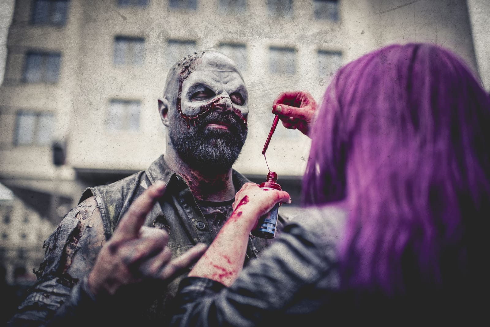 A zombie tilts his head back while a friend with purple hair drips fake blood onto his face from a bottle.