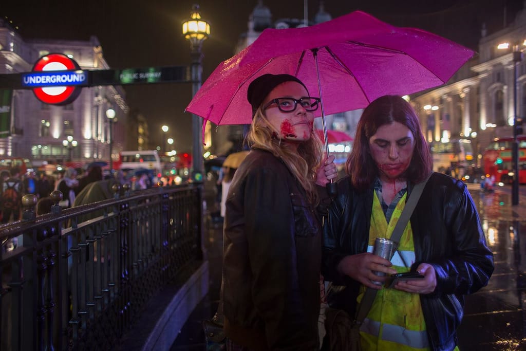 Two zombies shelter under a pink umbrella in the rain at Piccadilly Circus at night, beside a London Underground sign.