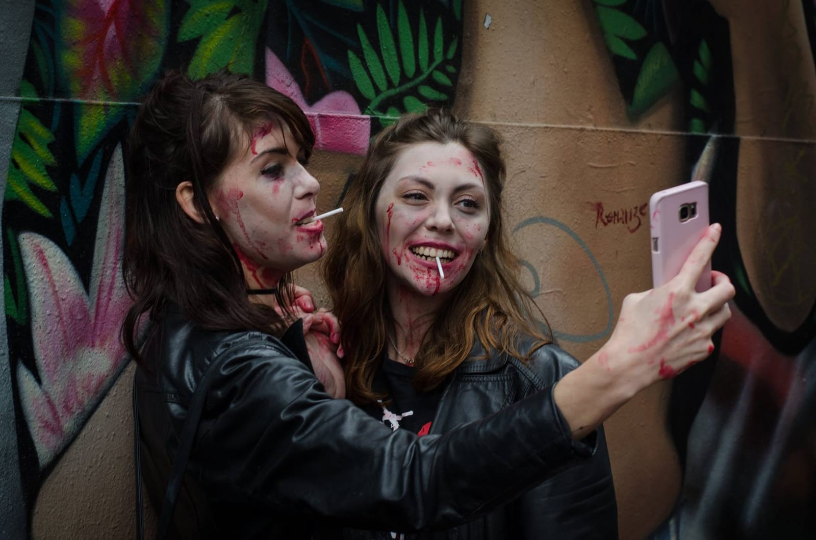 Two zombies with lollipops in their mouths take a selfie against a brightly painted graffiti wall.