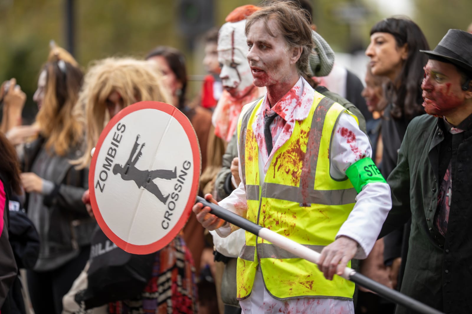 A steward in a blood-spattered high-vis vest holds a hand-painted “Zombies Crossing” lollipop sign while the walk waits behind him.