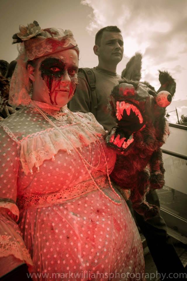 A zombie bride in a pink lace dress holds a snarling wolf puppet, another participant standing behind her.