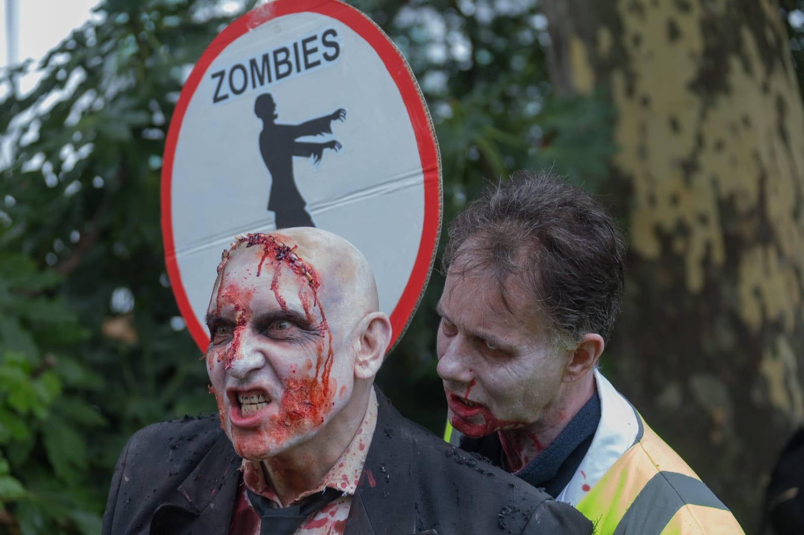 Two zombies grimace beneath a hand-painted “Zombies” crossing sign held above them.